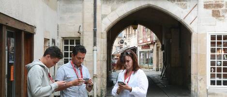 Three people standing in a cobblestone street with historic buildings, including an archway and a coat of arms.