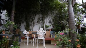Área de estar ao ar livre com cadeiras brancas e um banco, cercada por diversos vasos de plantas e árvores.