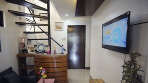 Uma área de recepção com uma mesa de madeira, uma tela de TV exibindo um preço e uma escada com corrimão de metal.