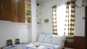 Quarto com guarda-roupa de madeira, cama com roupa de cama floral, mesa de cabeceira com livros e janela com cortinas estampadas.