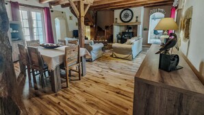 Un intérieur rustique avec des poutres apparentes, un coin repas avec des chaises en bois, un canapé et une cheminée surmontée d’une horloge.