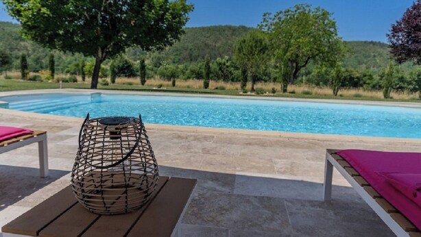Un espace aménagé au bord de la piscine, avec un fauteuil en osier et deux chaises longues, entouré d’un jardin paysager et d’arbres.