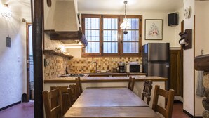 A kitchen with wooden cabinets, a tiled backsplash, a dining table with chairs, a refrigerator, and a window with wooden frames. - Apartment in the heart of the village (Puget-Théniers)