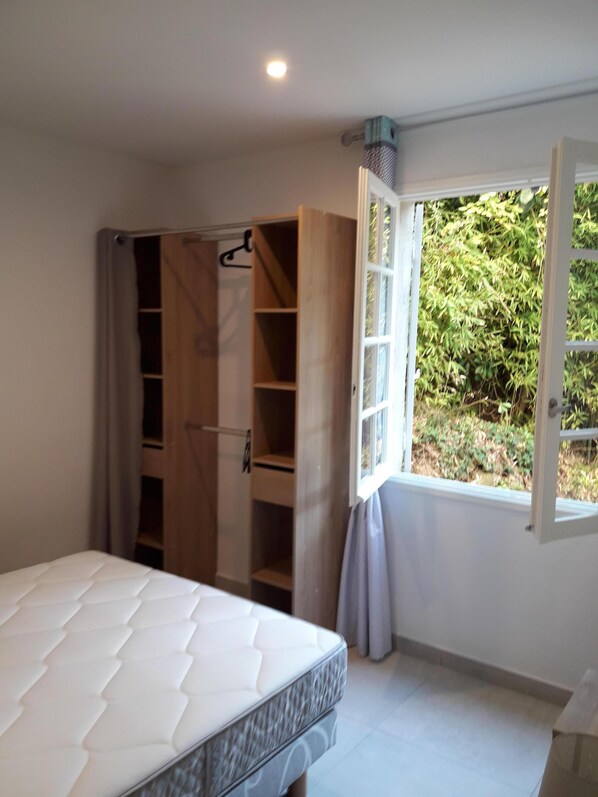 A bedroom with a bed, a wooden wardrobe, and a window with a view of greenery.