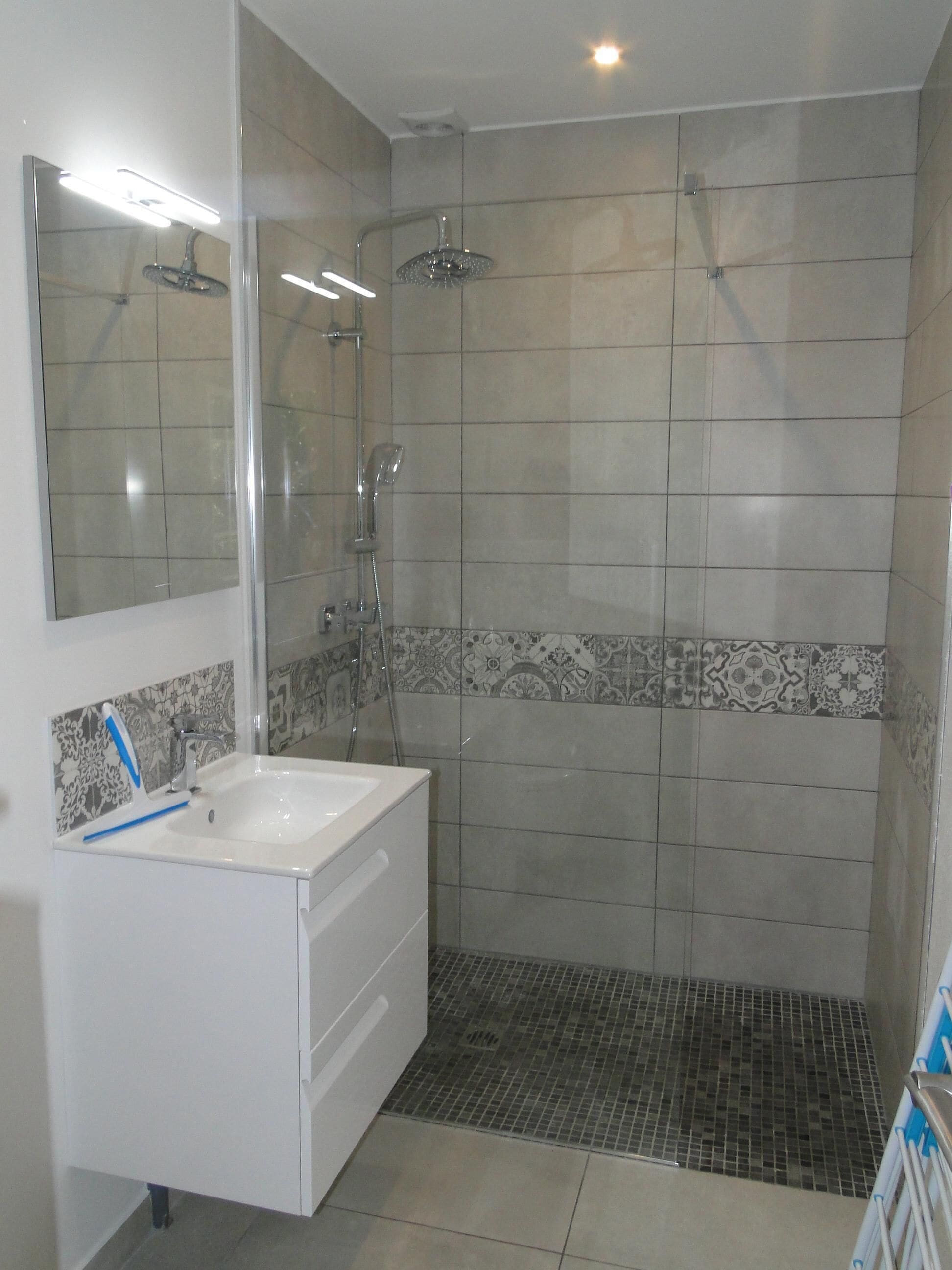 Un baño con tocador blanco, espejo, ducha con puerta de cristal y azulejos estampados.