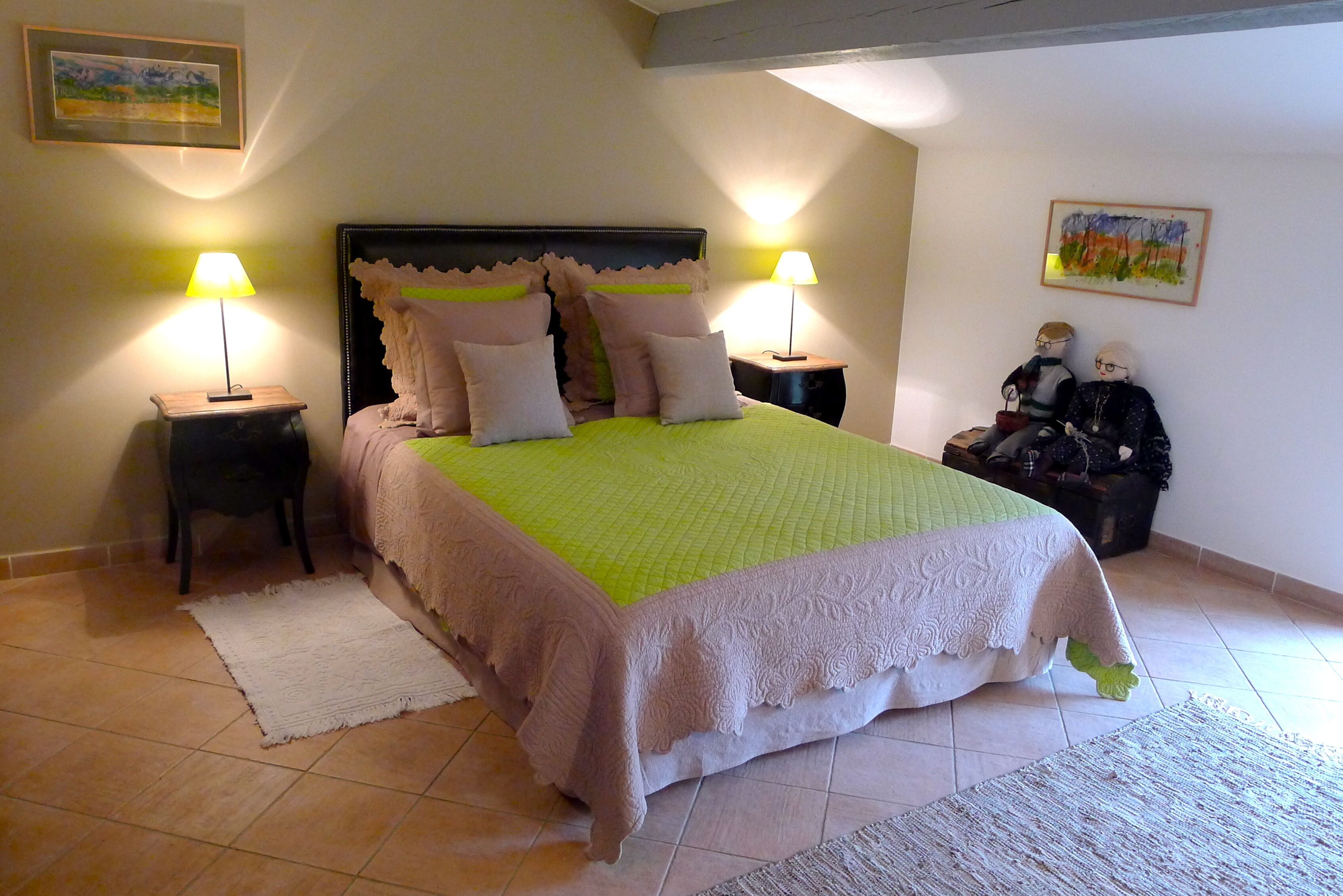 A neatly made bed with a green and white bedspread, flanked by two nightstands with lamps, in a room with tiled flooring and a painting on the wall.