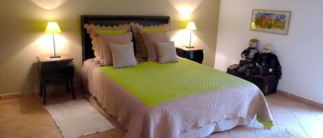 A neatly made bed with a green and white bedspread, flanked by two nightstands with lamps, in a room with tiled flooring and a painting on the wall.