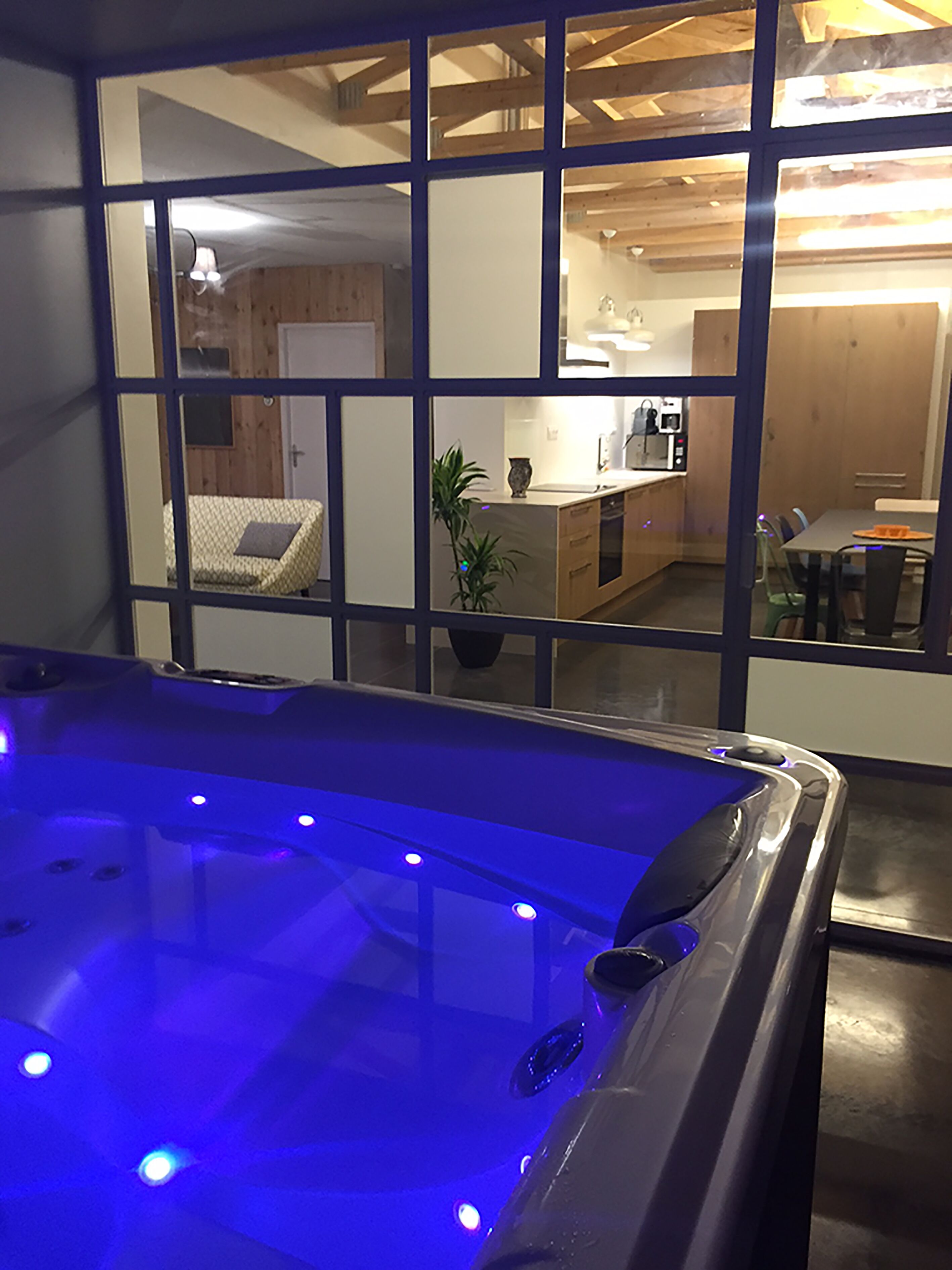 A modern bathroom with a glass-enclosed hot tub, wooden ceiling beams, and a visible kitchen area in the background.