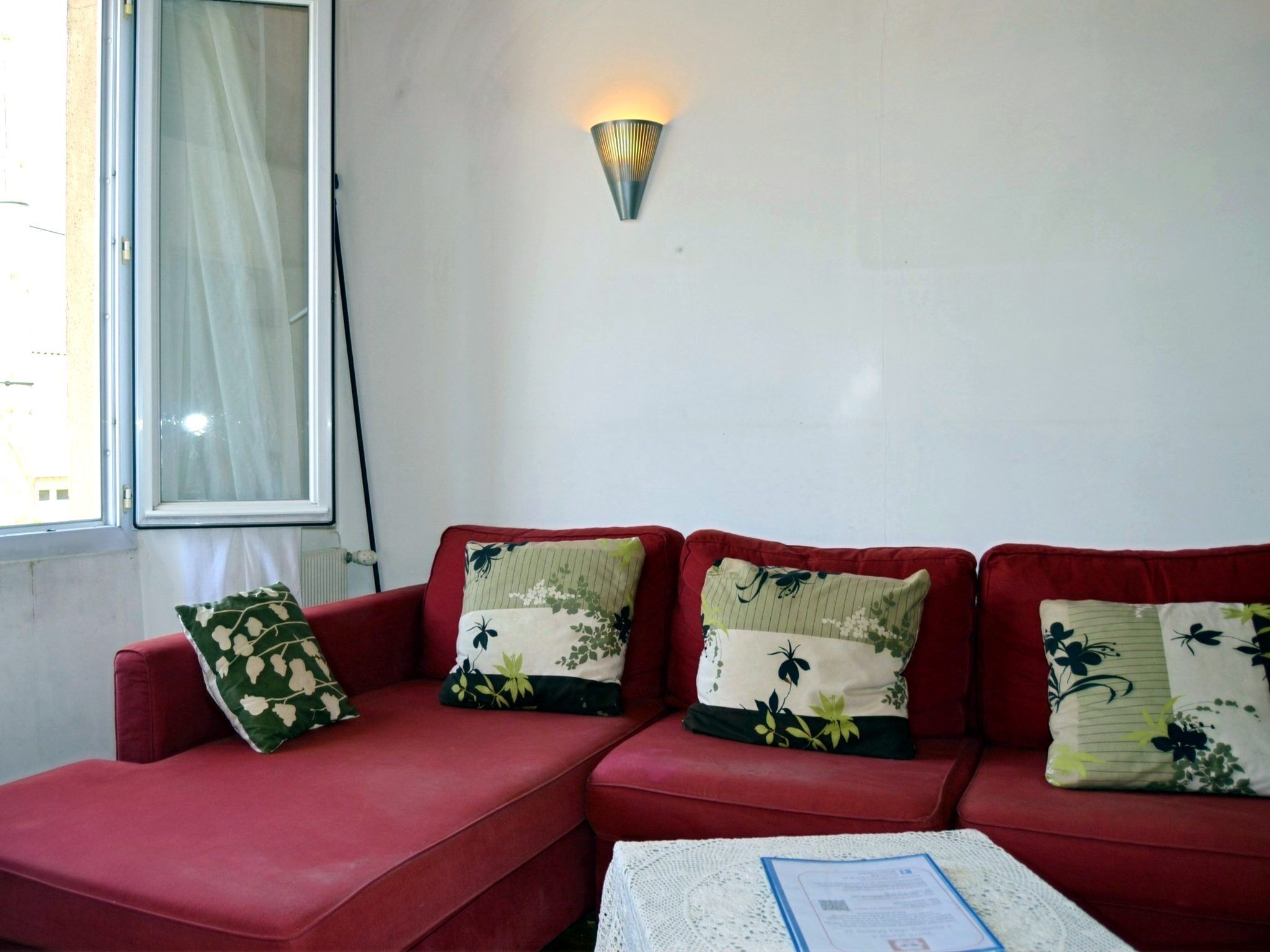 A red sofa with floral cushions, a white table with a document, and a wall-mounted light fixture.