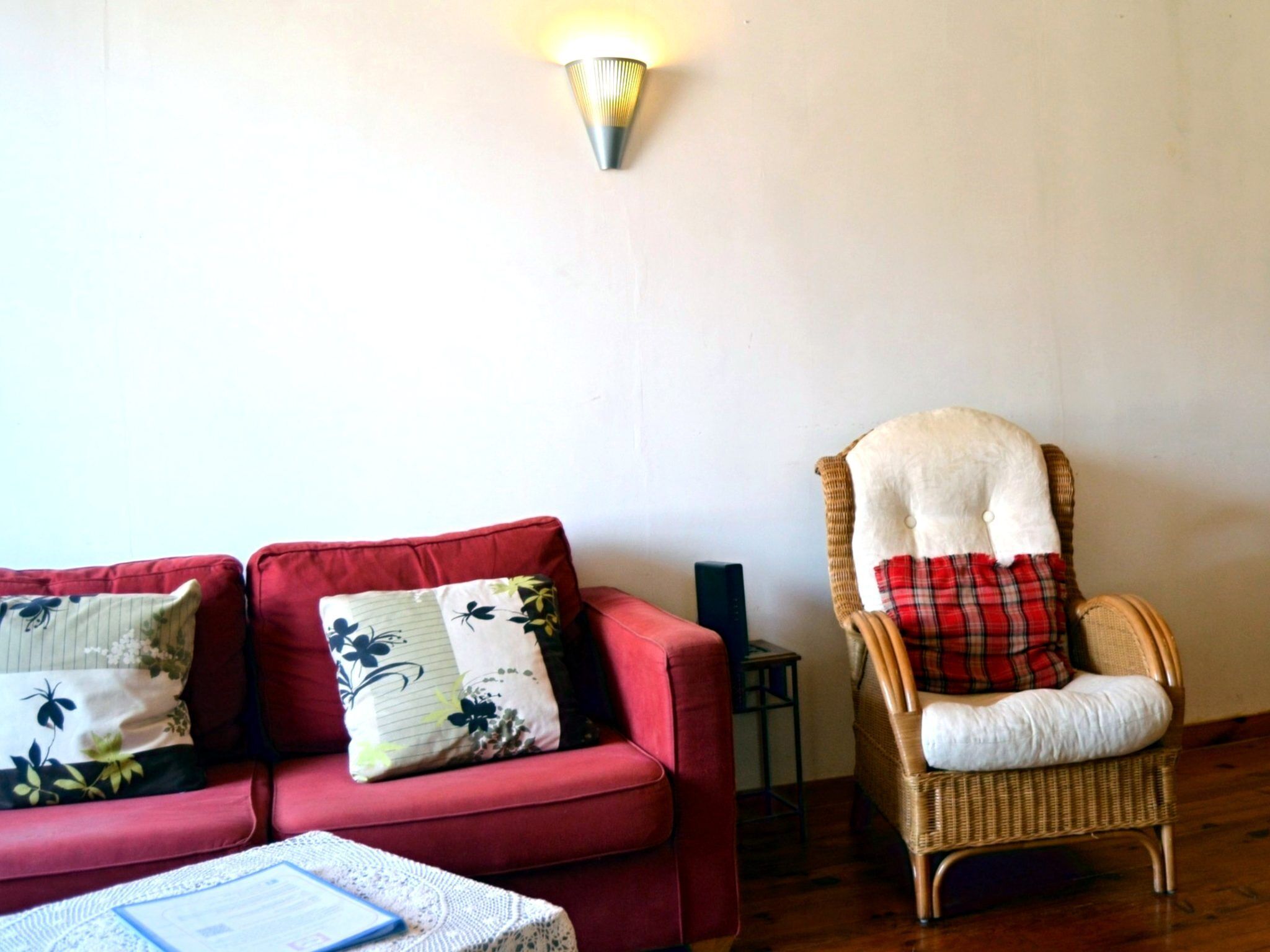 A red sofa with floral cushions, a wicker chair with a plaid cushion, and a wall-mounted light fixture.