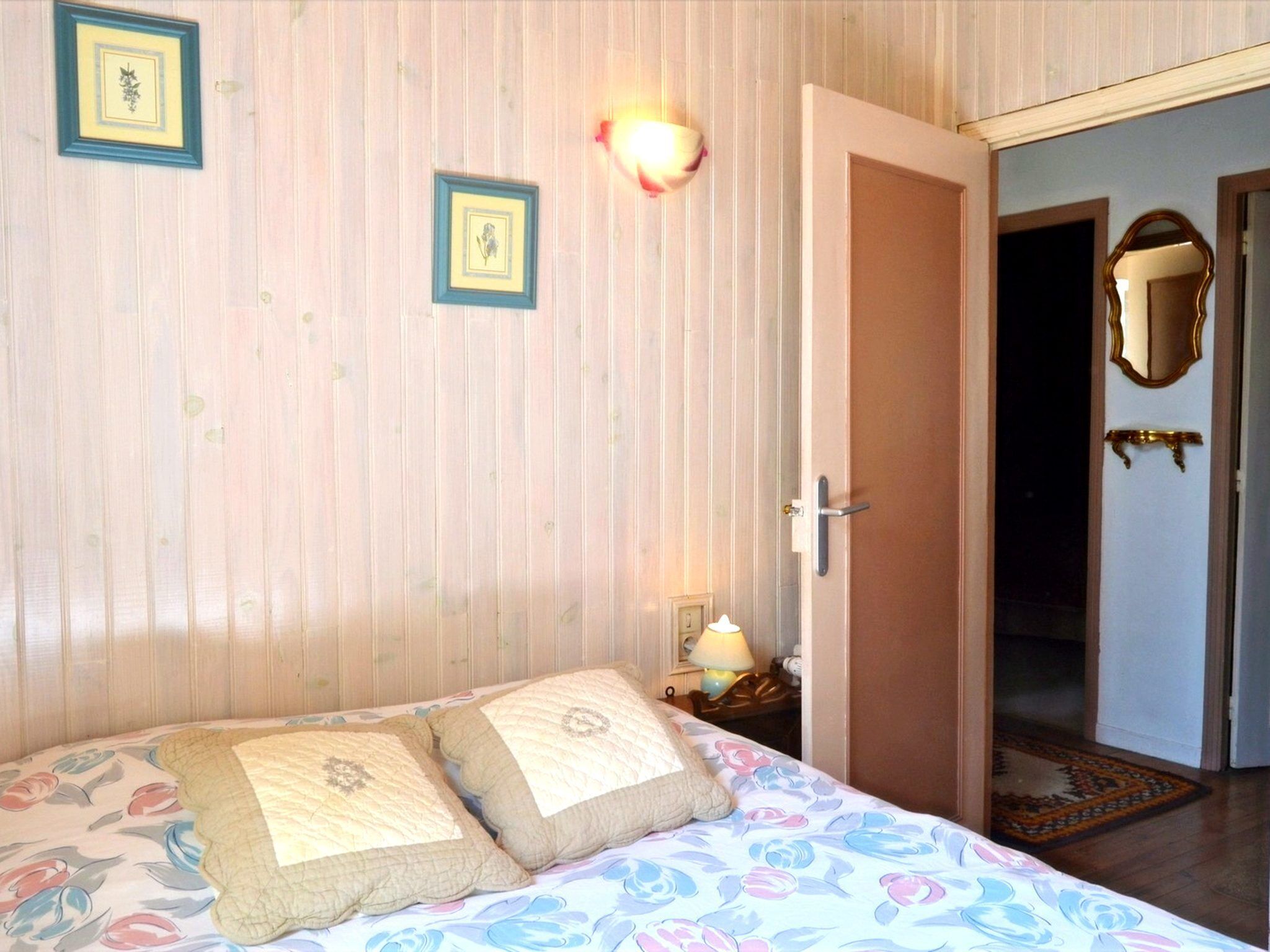 Een slaapkamer met een bed, twee kussens, een nachtkastje, een lamp, een deur en een spiegel.