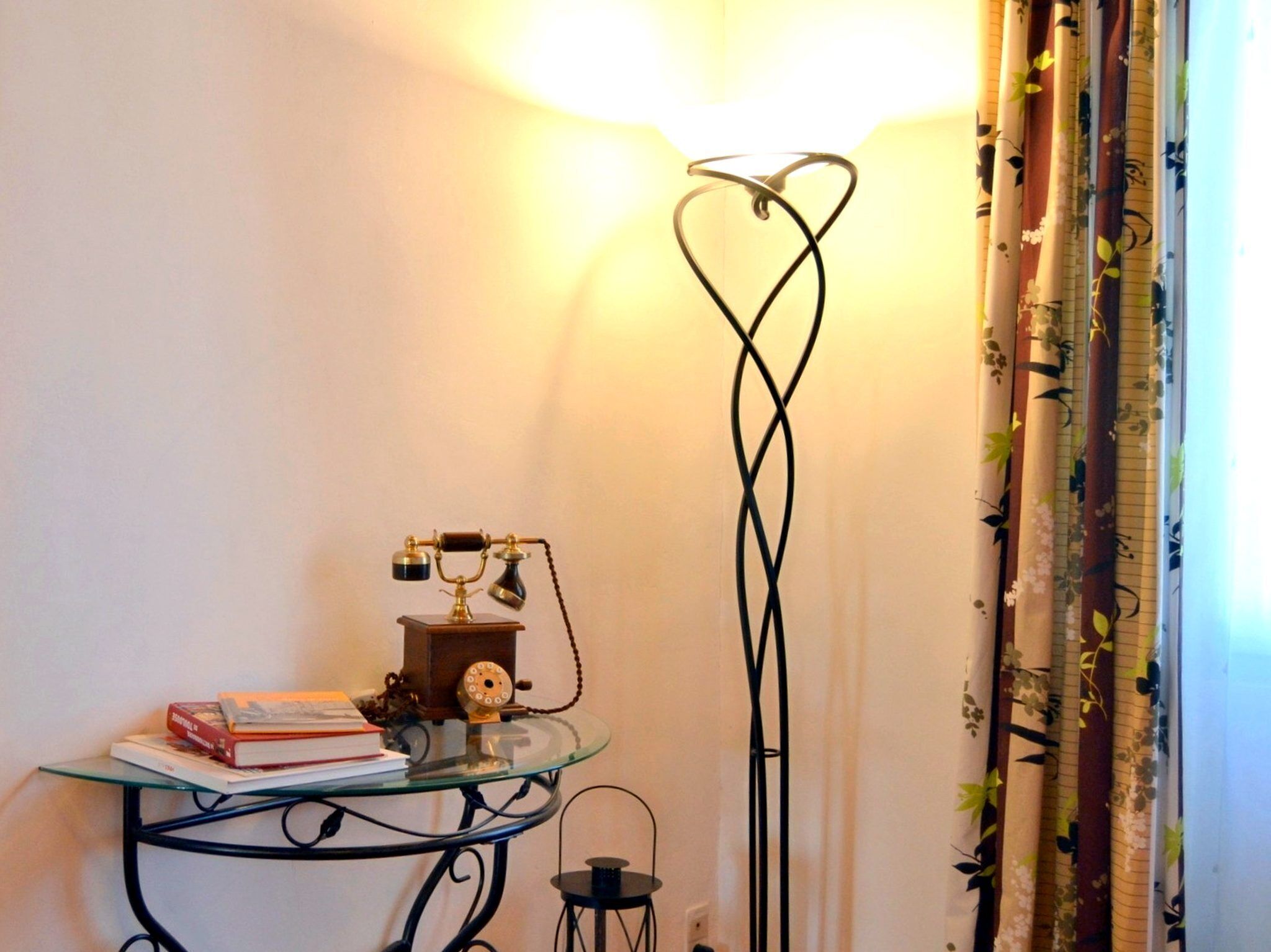 A corner of a room with a glass table holding books and a vintage telephone, a floor lamp with a heart-shaped base, and curtains with a floral pattern.
