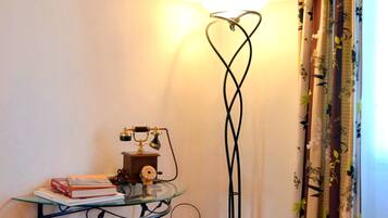 A corner of a room with a glass table holding books and a vintage telephone, a floor lamp with a heart-shaped base, and curtains with a floral pattern.