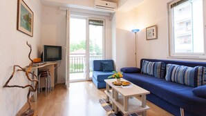 A living room with a blue sofa, a small table with a vase, a TV on a stand, and a balcony door. - Exclusive Apartment Tiffany Ohrid (Ohrid)