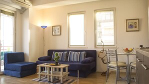 A living room with a blue sofa, a small table with a plant, and a dining area with metal chairs and a fruit bowl. - Exclusive Apartment Tiffany Ohrid (Ohrid)