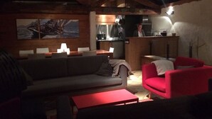 Een gezellige zithoek met een bank, een rode fauteuil en een eettafel. Achtergrond toont een keuken waar iemand kookt.