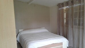 Una cama bien tendida con estructura de madera y ropa blanca en una habitación con paredes color beige y un ventanal con cortinas traslúcidas.