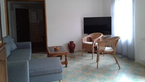 Una sala de estar con sofá, televisor, sillones de mimbre, una mesita y una ventana con cortinas.