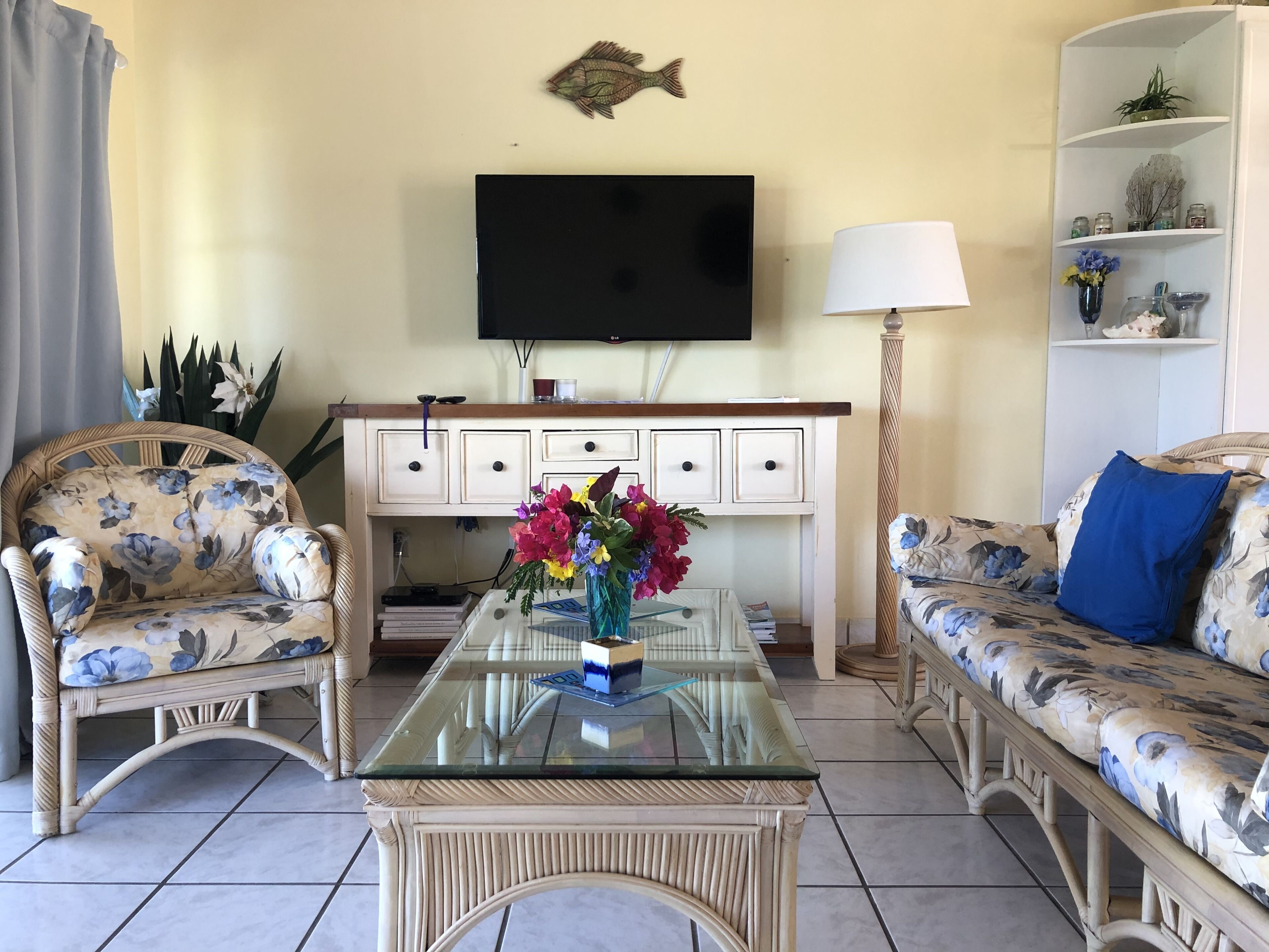 A living room with a glass-top coffee table, wicker furniture, a flat-screen TV, and a bookshelf with decorative items.