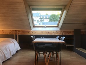 Ein kleiner, gemütlicher Raum mit schrägem Dach und einem Oberlicht. Er verfügt über einen Holztisch mit Stühlen, ein Bett mit weißer Decke und eine Sitzbank an der Wand.