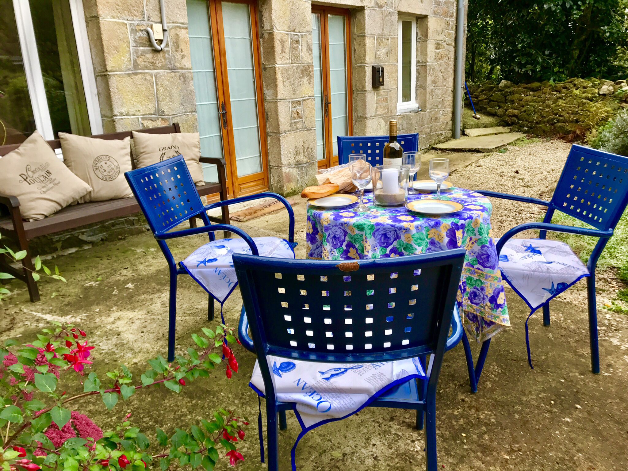 Un área de estar al aire libre con sillas azules y una mesa lista para comer, rodeada de muros de piedra y vegetación.