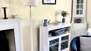 Ein Wohnzimmer mit weißem Kamin, einer stehenden Stehlampe, einem Schrank mit Geschirr und einem blauen Sessel.