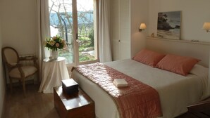 Ett sovrum med en säng, en stol, ett bord med en vas med blommor, en lampa och en tavla på väggen.