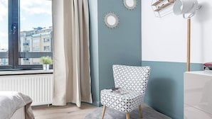 Une chambre à coucher moderne donnant sur des immeubles par une fenêtre, avec un fauteuil à motifs, un radiateur et une commode blanche.