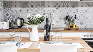 Une cuisine moderne avec une table à manger dressée pour quatre personnes, un pichet blanc orné de fleurs, une bouteille d’huile d’olive et un panier de fruits posé sur le comptoir.