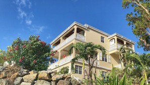 A multi-story building with balconies, surrounded by rocks and tropical plants. - 
2 BEDROOMS WITH OCEAN VIEW AND GARDEN VIEW IN JOST VAN DYKE – SUNRISE (Jost Van Dyke)
