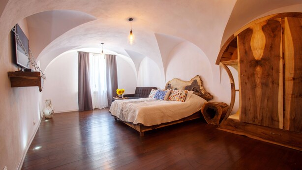 Een slaapkamer met een houten bed, een groot raam met gordijnen en een bijzonder gewelfd plafond.