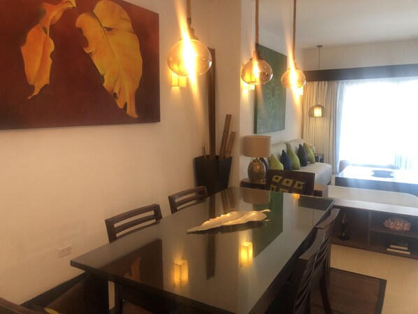 A modern dining area with a glass table, chairs, and pendant lights. There is a painting on the wall and a sofa with pillows in the background.