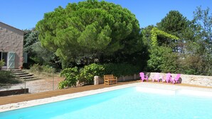 A swimming pool with a wooden deck, surrounded by a stone wall and various plants. There are wooden chairs and a bench near the pool. - Vacation rental for 3 people, 1 bedroom, pool, nature and peace and quiet guaranteed (Laurac)