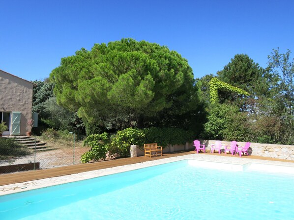 Une piscine dotée d’une terrasse en bois, entourée d’un mur de pierre et agrémentée de diverses plantes. On y trouve des chaises et un banc en bois, situés à proximité de la piscine.