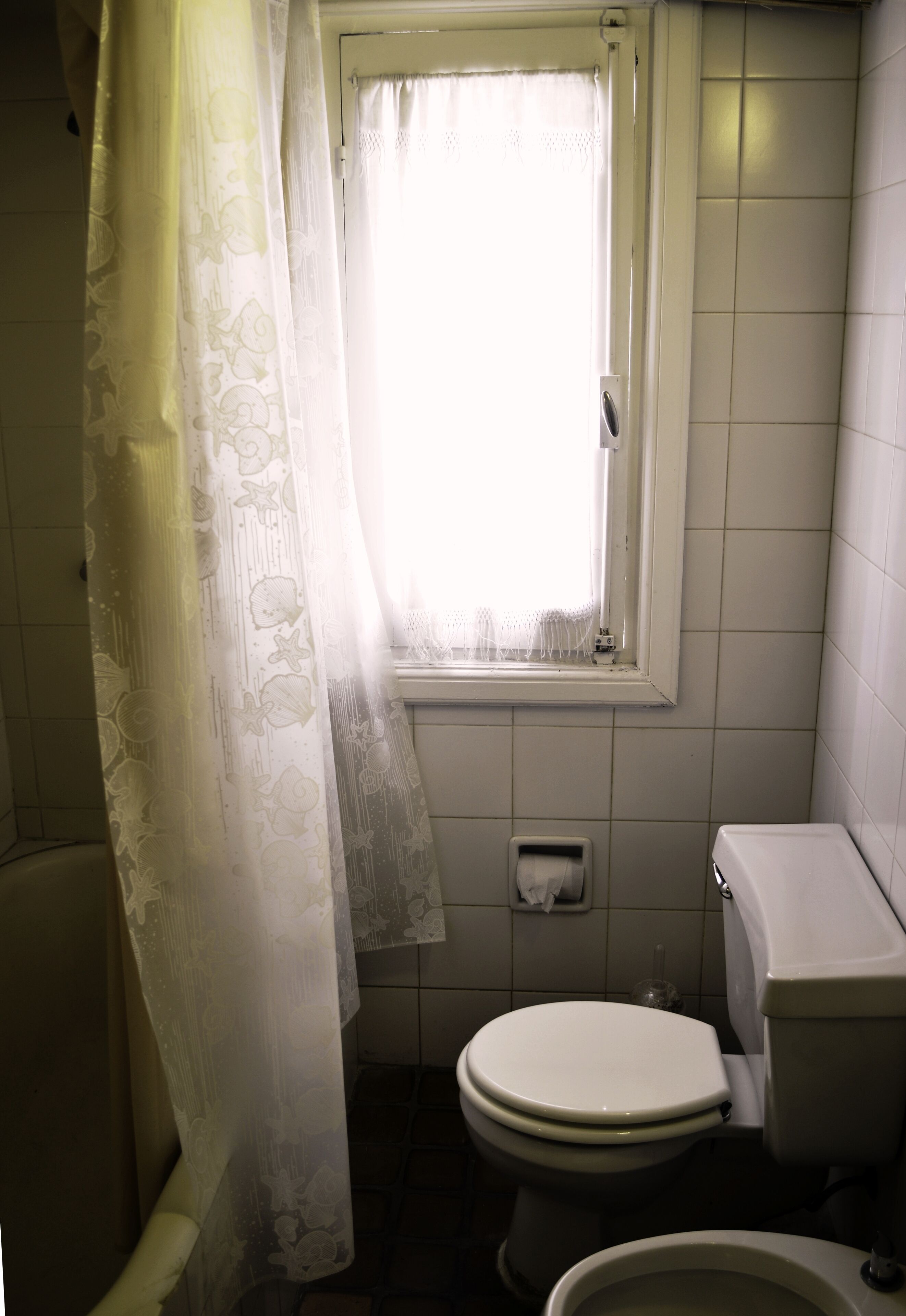 Een kleine badkamer met een toilet, een raam met gordijn en een betegelde muur.