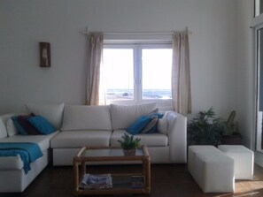 Un salon avec un canapé blanc, une petite table basse en bois et une vue sur la mer par une grande fenêtre.