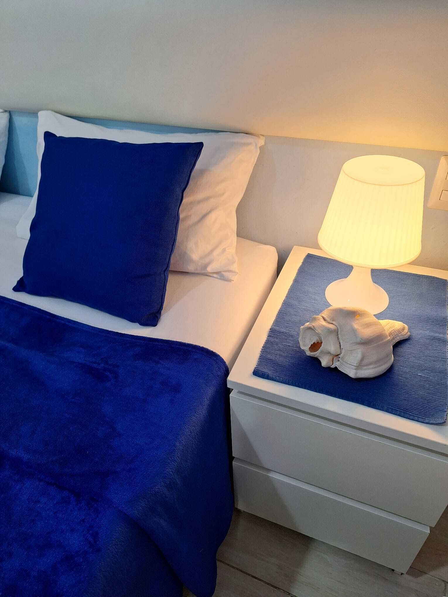 A bed with a blue blanket and pillows, a bedside lamp, and a small white object on a blue mat.