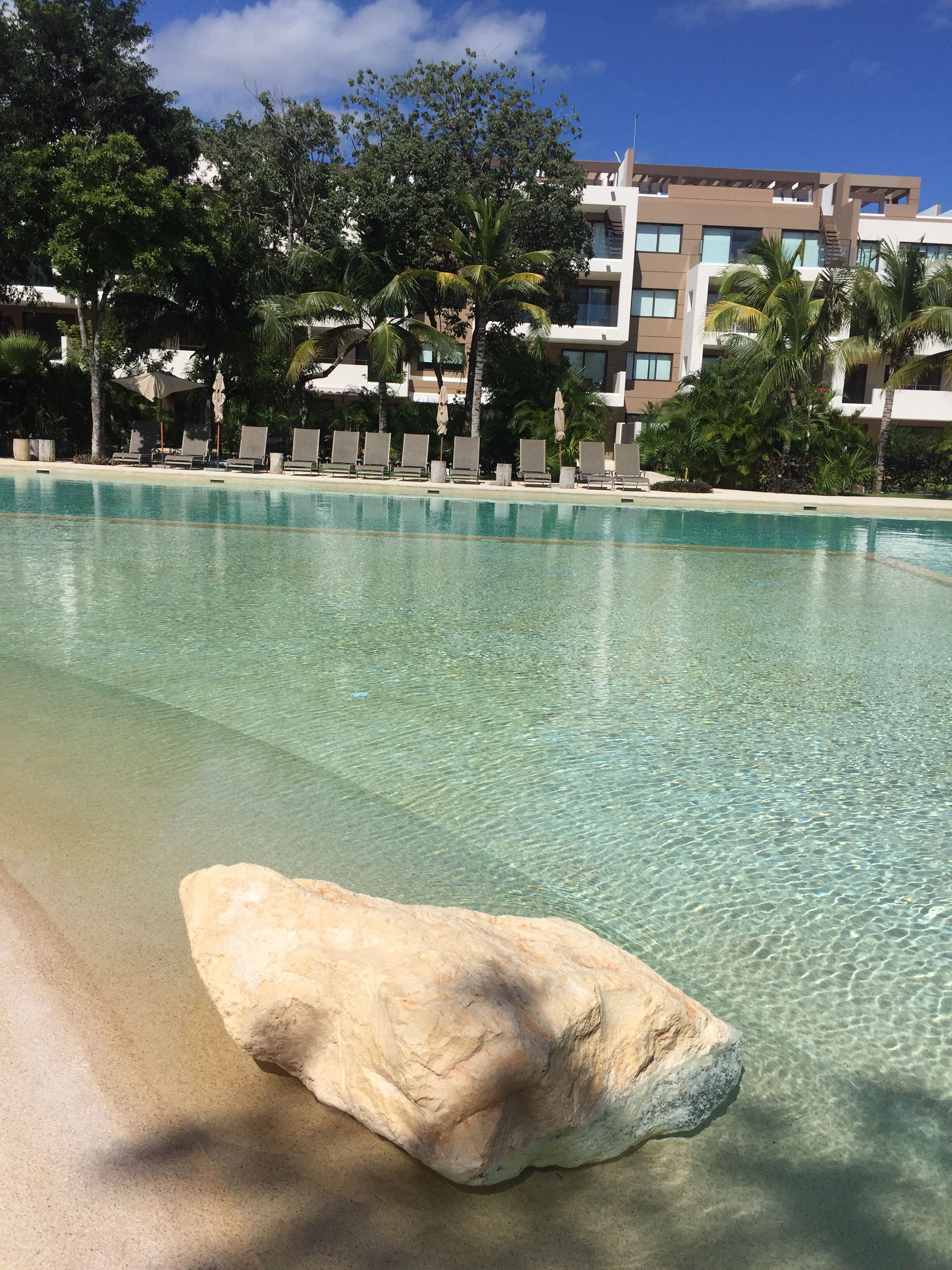 A clear pool with a large rock in the foreground, surrounded by lounge chairs and umbrellas, with a modern building in the background.