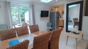 A dining area with wicker chairs and a table, a kitchen in the background, and a TV mounted on the wall. - Les Muguets Residence - Ground-Floor Apartment (Villers-sur-Mer)