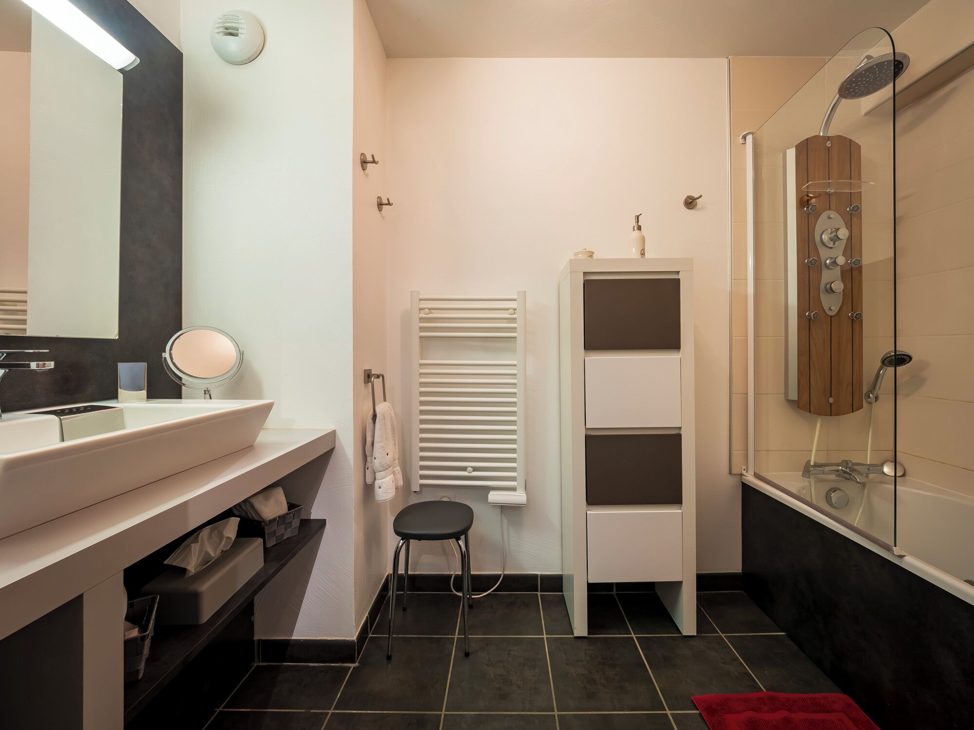 Un bagno moderno con un mobile lavabo bianco, uno specchio rotondo, un porta asciugamani, un piccolo sgabello, un termosifone e una cabina doccia in vetro.