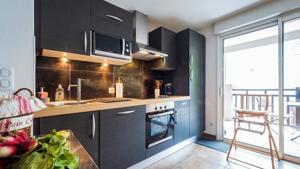 Een moderne keuken met donkere kastjes, een ingebouwde magnetron en een oven. Er is een houten aanrecht met een mand vol groenten en een fles olijfolie.