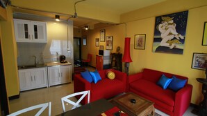 Un espace de vie compact comprenant un coin cuisine, des canapés rouges avec des coussins bleus, une table à manger et une lampe rouge.