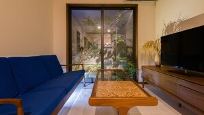 A living room with a sofa, a coffee table, a TV, and a sliding glass door leading to a balcony with plants.