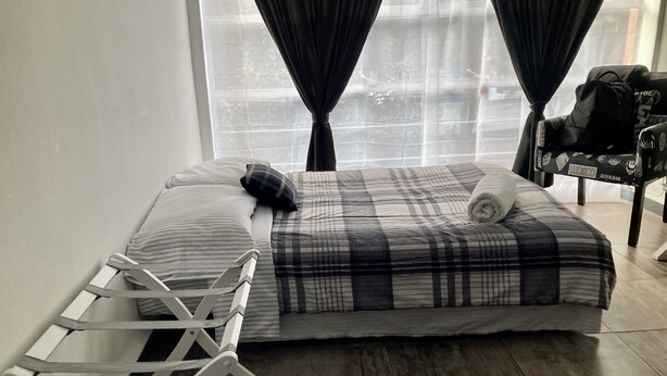 A neatly made bed with a plaid bedspread and pillows, a white metal crib, and a chair with a black bag.