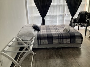 Una cama bien tendida con una colcha escocesa y almohadas, una cuna de metal blanco y una silla con un bolso negro.