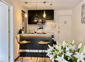Eine moderne Küche mit schwarzer Arbeitsplatte, Edelstahlgeräten und Pendelleuchten. Zwei Barhocker und eine Vase mit weißen Lilien sind ebenfalls vorhanden.