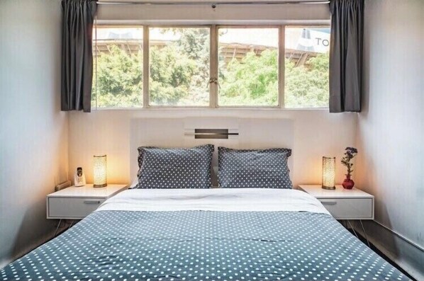 Een slaapkamer met een bed, twee nachtkastjes, een raam met gordijnen en uitzicht op groen.