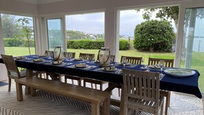Uma sala de jantar com uma mesa comprida posta para seis pessoas, janelas amplas e vista para uma paisagem verde.
