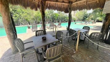 A covered outdoor seating area with tables and chairs, overlooking a pool and lush greenery.
