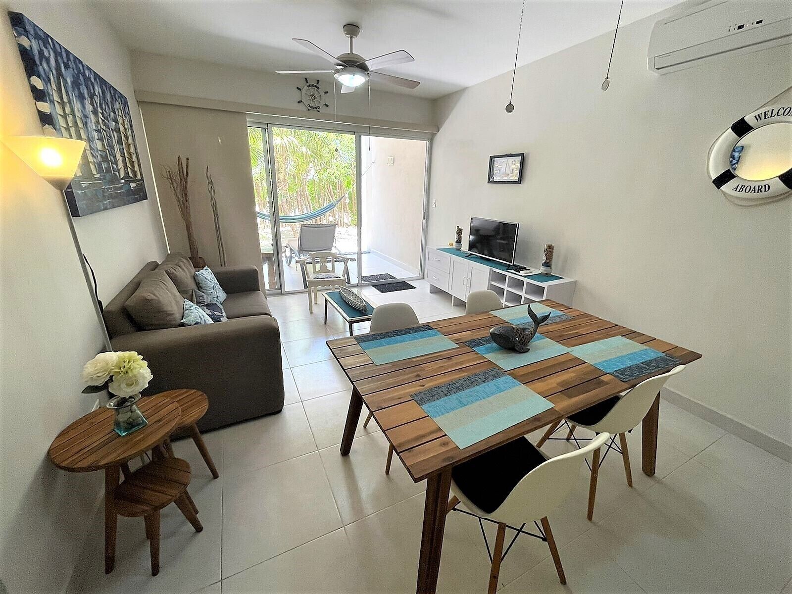 A living room with a sofa, a dining table with chairs, a TV, and a hammock outside the sliding glass door.
